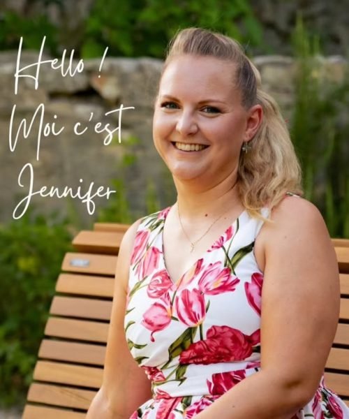 Photo de Jennifer, décoratrice de mariage à Aix-En-Provence, blonde, les cheveux attachée, souriante et assise sur un banc dans une jolie robe blanche à fleurs rouges